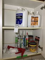 Shelf with opened cupboard containing tubes of caulking, the red and white caulking tool, several solvent and oil containers