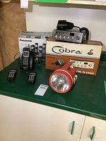 Overview of the Cobra CB radio on its box with microphone, Panasonic cordless phone set in box with base and handsets, and the large red flashlight.