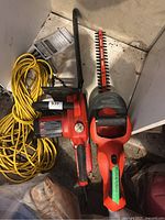 View of Black and Decker cordless hedger trimmer without battery, electric Homelite chainsaw and yellow extension cord laid on floor
