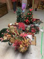Photo showing multiple Christmas decorations including a small decorated artificial tree, floral arrangements, wrapped boxes, and other holiday decor items grouped together on the floor.
