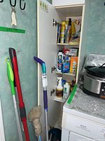 Open cupboard filled with assorted cleaning liquids and cloths, mop handles leaning against wall