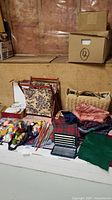 Wide view of the collection showing tapestry sewing bag, woven bag, fabric remnants, knitting needles, and small sewing box on the floor with other items spread out