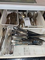 A drawer containing three plastic baskets filled with a collection of metal spoons, forks, and knives, including butter knives and serving utensils.