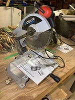 Side view of Delta Shopmaster compound miter saw on workbench with user manual and hardware bag on base.