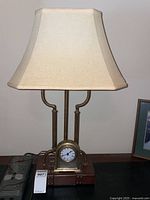 Full view of the desk lamp showing the beige shade, twin metal rods, faux briefcase styled base, and embedded clock at center front.