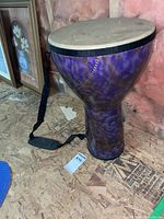 Full view of the purple and gold doumbek drum with shoulder strap sitting on a plywood surface.