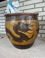Front view of round yellow-brown glazed planter pot with dark brown raised dragon motif and plastic bag of faux flowers inside