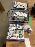 Dremel rotary tool in gray case with manuals, power cord visible, and accessory kit and small sealed accessory pack on table.