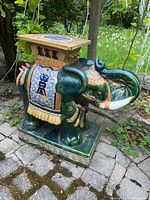 Front-side view of painted elephant stand on stone pavers, showing trunk raised, tassels on ear and body, square top
