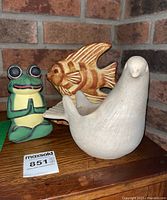 Photo showing all three figures: dove flower pot, fish figurine, and frog figurine on wooden surface in front of brick background.