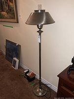 Full view of the metal floor lamp showing the conical metal shade with clear glass chimney, brushed pole, and round base against a wall.