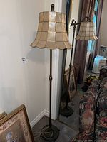 Full view of the floor lamp standing against a wall near a mirror and floral furniture, showing the solid brass base, vertical rod, and beige shade.
