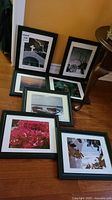 Seven black framed and matted photos arranged on wooden floor leaning against wall and small table