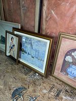 Photo showing all four framed prints leaning against a wall in a basement area, displaying different styles and themes: Royal portrait, sailboats, floral, and train print on the far left.