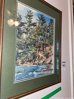 Framed watercolour painting showing a scenic landscape with trees, a rocky cliff, water, and a cabin.
