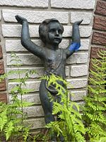 Front angle of cherub garden statue with arms raised, set against light-colored brick and surrounded by green ferns.