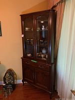Front corner angle view of dark wood corner china cabinet with glass doors, lighted upper section with two glass shelves, drawer and lower double door storage.