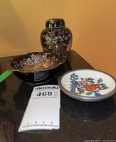 Front view of the black Japanese ginger jar with floral patterns, accompanied by one black floral china dish and one colorful round floral dish.