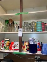 Two shelves showing an assortment of colorful coffee mugs with various designs and glass jars and glasses.