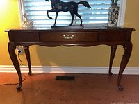 Front view of wooden hallway table with decorative items on top, showing curved cabriole legs and ornate brass handle on drawer.