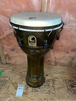 Full view of the Toca djembe drum showing the white drumhead, metal tuning rods, and gold and brown base.
