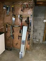 Full view of basement wall showing various hand tools hung including clamps, saws, level, pipe wrench, and track panels leaning against wall.