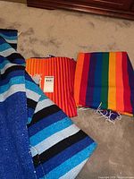 Photo of three folded blankets showing the rainbow striped one, a red and orange striped one, and part of the blue striped blanket.