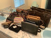 Overview of various handbags arranged on a bathroom sink counter including leather bags, woven bags and colorful Nepalese style bag