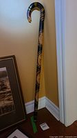 Full view of the handcarved walking stick showing black base color with multiple circular Aboriginal styled carvings and painted designs along the shaft, curved handle shaped like a bird's head.