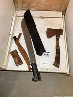 Four vintage tools displayed on white surface including hatchet with sheath, machete with sheath, filet tool, and pocket tool in leather holders.