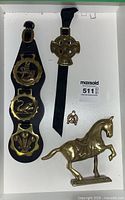 Photo shows collection of vintage brass horse brasses on black leather straps and a brass horse figurine, all placed inside a white box.