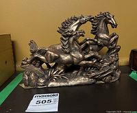 Frontal view of bronze-colored cast figurine depicting three galloping wild horses on a scenic base with foliage and rocks.