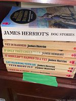 Stack of 7 soft cover James Herriot books laid horizontally, showing the full cover and spines for identification - James Herriot's Dog Stories is on top.