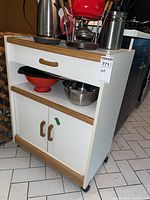 Front view of white kitchen cart with wood-toned trim, showing top drawer, open middle shelf with kitchen items, and closed two-door cabinet below.