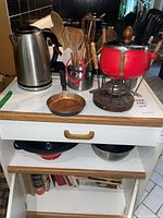 Long shot showing assortment of kitchen items on table: red fondue pot with wooden handles, electric kettle, two containers with utensils, copper frying pan, metal bowls, and cookbooks on lower shelf.