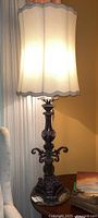 Front view of lamp showing beige scalloped fabric shade and ornate metal base with scrollwork, illuminated to show light diffusion.