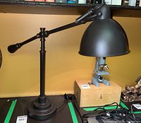 Black adjustable desk lamp with wired power cord on a black surface, with a microscope and books in background.