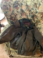 Brown leather jacket laid on floral patterned chair demonstrating worn condition and overall design.