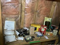 Wide shot of various security and lighting equipment on a wooden shelf, showing items including multiple smoke alarms in boxes, keypads, thermostat unit, and accessories.