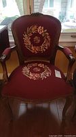 Front view of burgundy needlepoint armchair with wooden frame, showing floral wreath embroidery on seat and backrest.