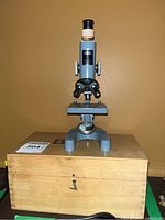 Front view of the Lumex microscope placed on top of the wooden box showing eyepiece, rotating objective lenses, focus knobs, and stage.