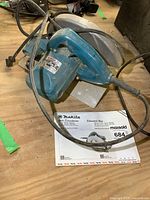 Makita circular saw photographed on wooden surface with instruction manual underneath showing branding and model information.