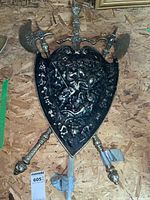 Front view showing replica black medieval decorative shield with embossed knight battle scene and three attached weapons (axe, sword, mace).