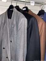 Three men's jackets on hangers showing a gray wool patterned jacket, a black jacket, and a brown suit jacket.