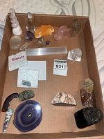 Photo showing various mineral and crystal samples arranged in a cardboard box including selenite wand, quartz crystals, amethyst, agate slice, kyanite in labeled bag, and other mineral rocks.