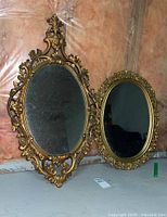 Two ornate oval gilded mirrors side by side on floor against a plastic-covered wall, showing full frames and aged mirror surfaces.