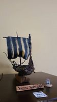Full view of the Viking war ship model showing the blue and white striped fabric sail, carved wooden hull, mounted on a wooden block base with a detached piece and a wooden plaque reading 'Viking War Ship'.