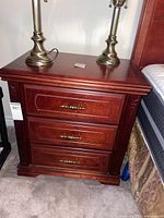 Photo showing one of the wooden night tables next to bed, dimensions 28x16x28, three drawers with brass handles, with two lamps on top
