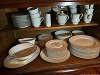 Full view of the Nitto Cluny Lace china plates, cups, platters, and serving dishes arranged inside a wooden cabinet, showing different sizes and types of plates and cups stacked neatly.