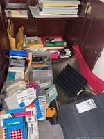 Open wooden cabinet displaying organized office supplies including notebooks, envelopes, pens, clips, files, a Dymo label maker, and a black computer case.